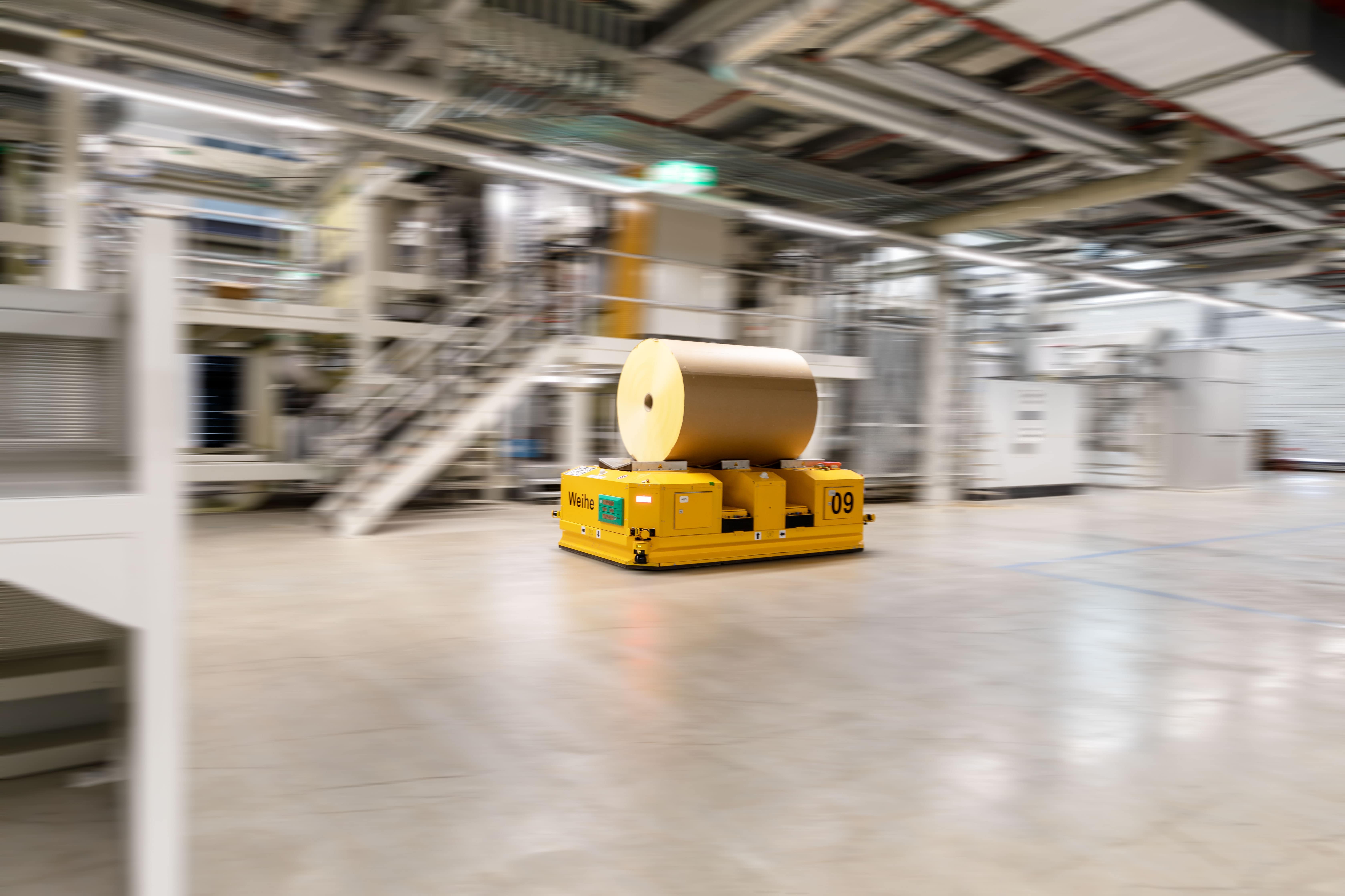A yellow automated transport cart moves with a large roll of paper in a modern factory hall.