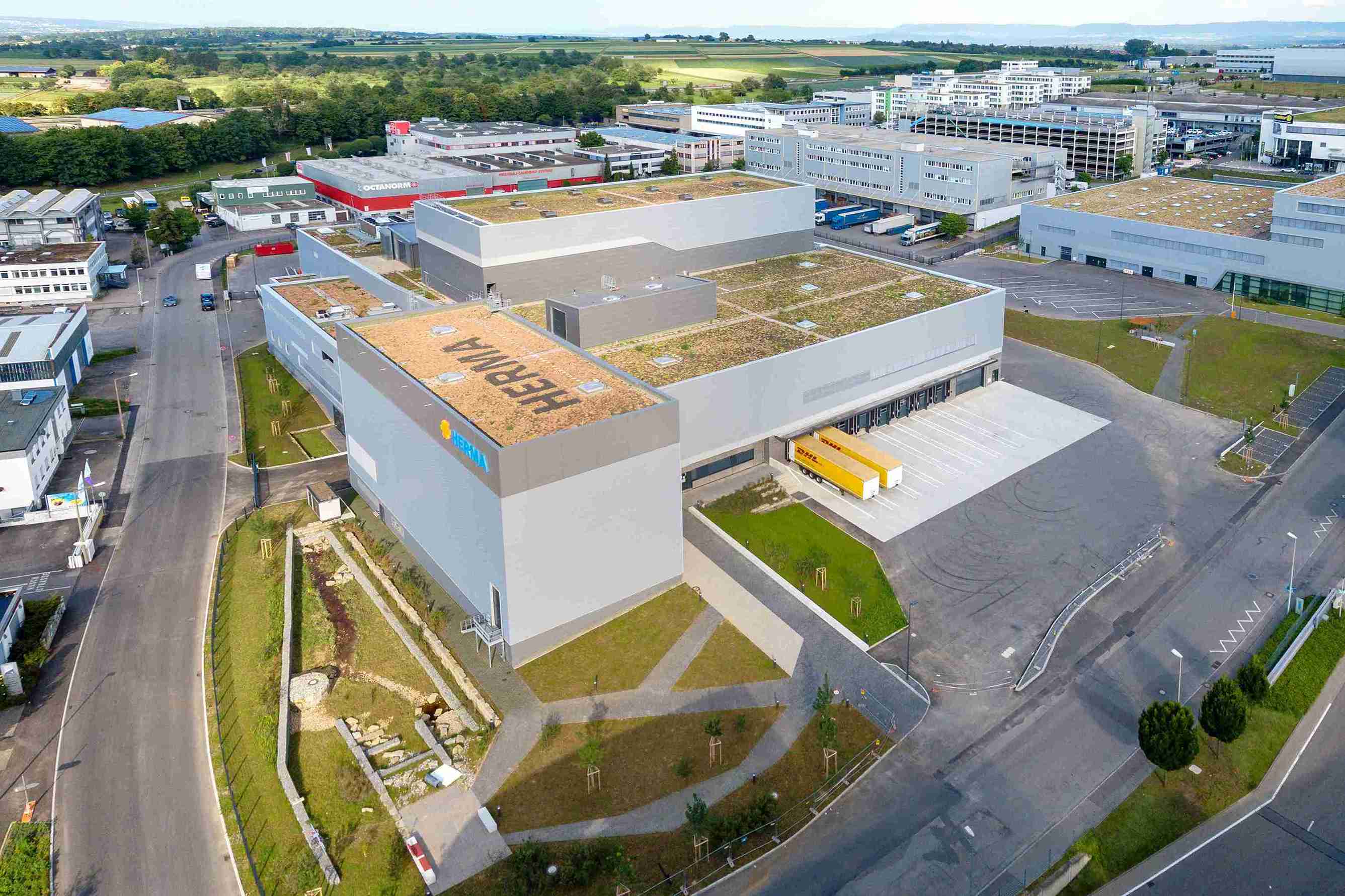 Aerial view of a modern industrial building with a green roof and surrounding parking lots, part of a larger commercial area.