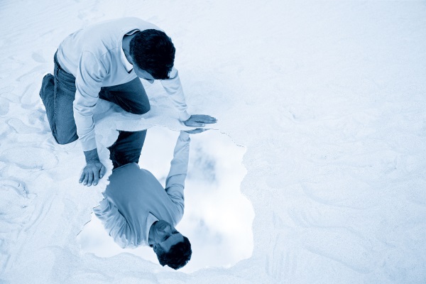 A man kneels in the sand and gazes at his reflection in a small puddle of water.