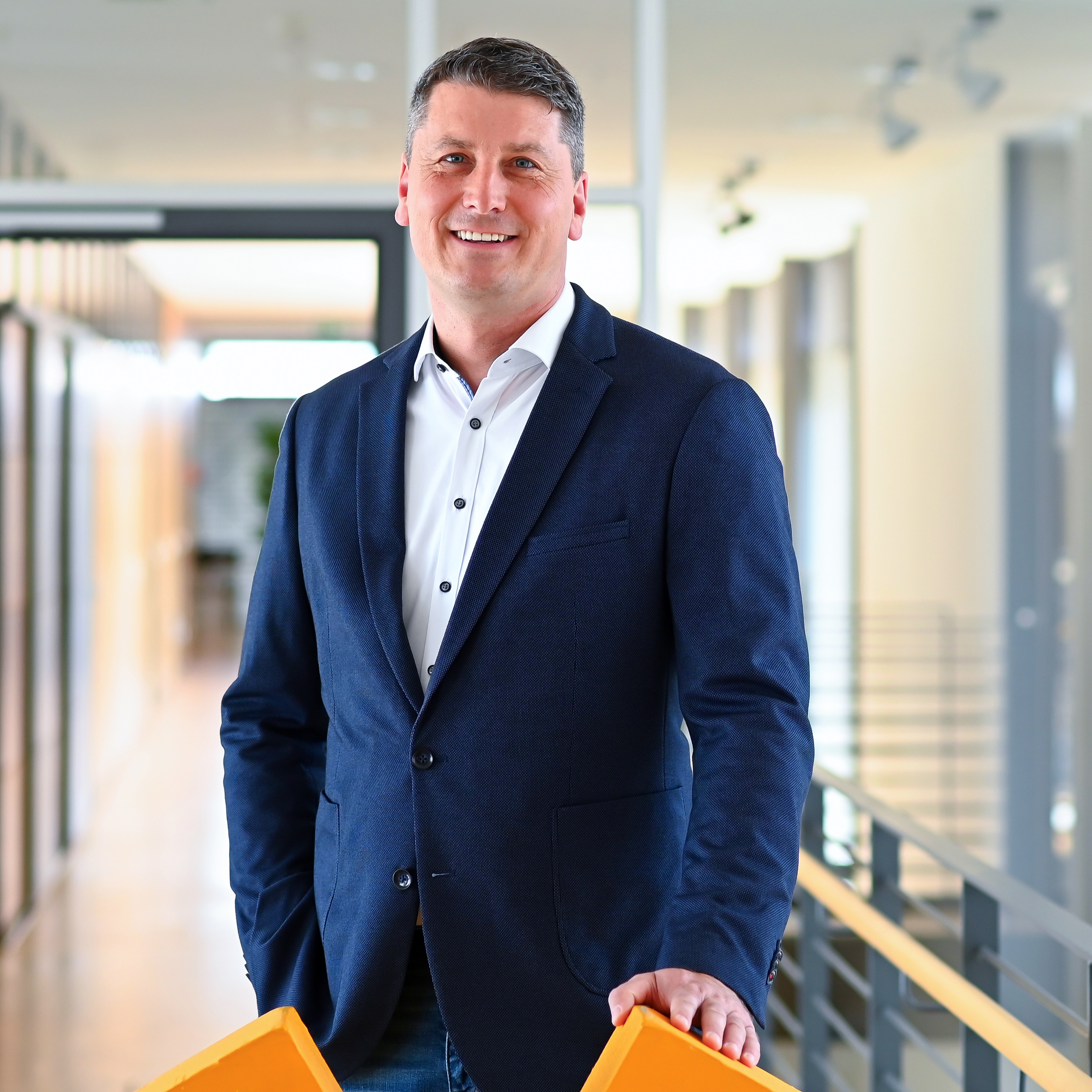 A man in a blue blazer stands in a modern office hallway, holding two orange objects in his hand.