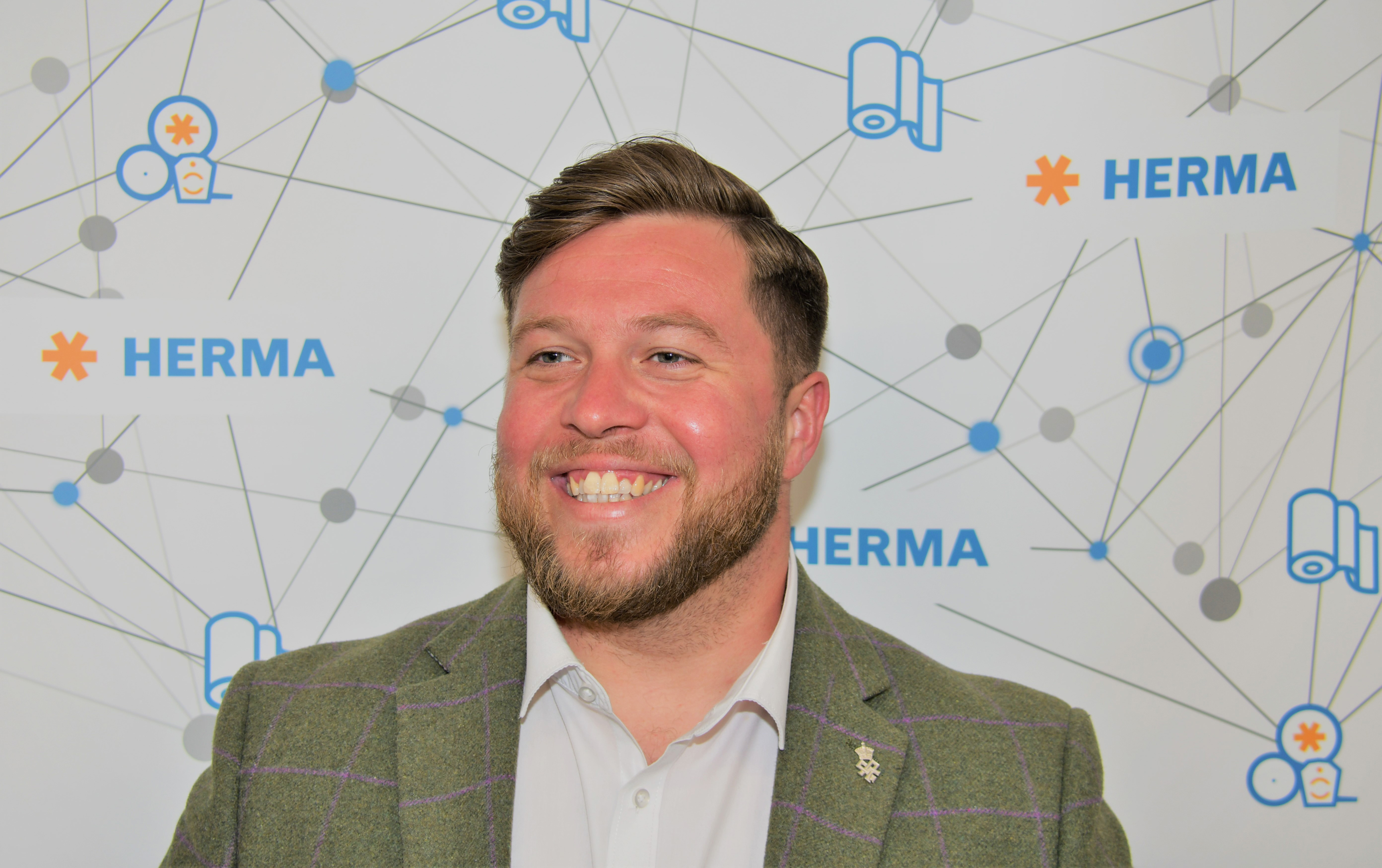 A man with short hair and a beard smiles against a background featuring the HERMA logo and graphic elements.