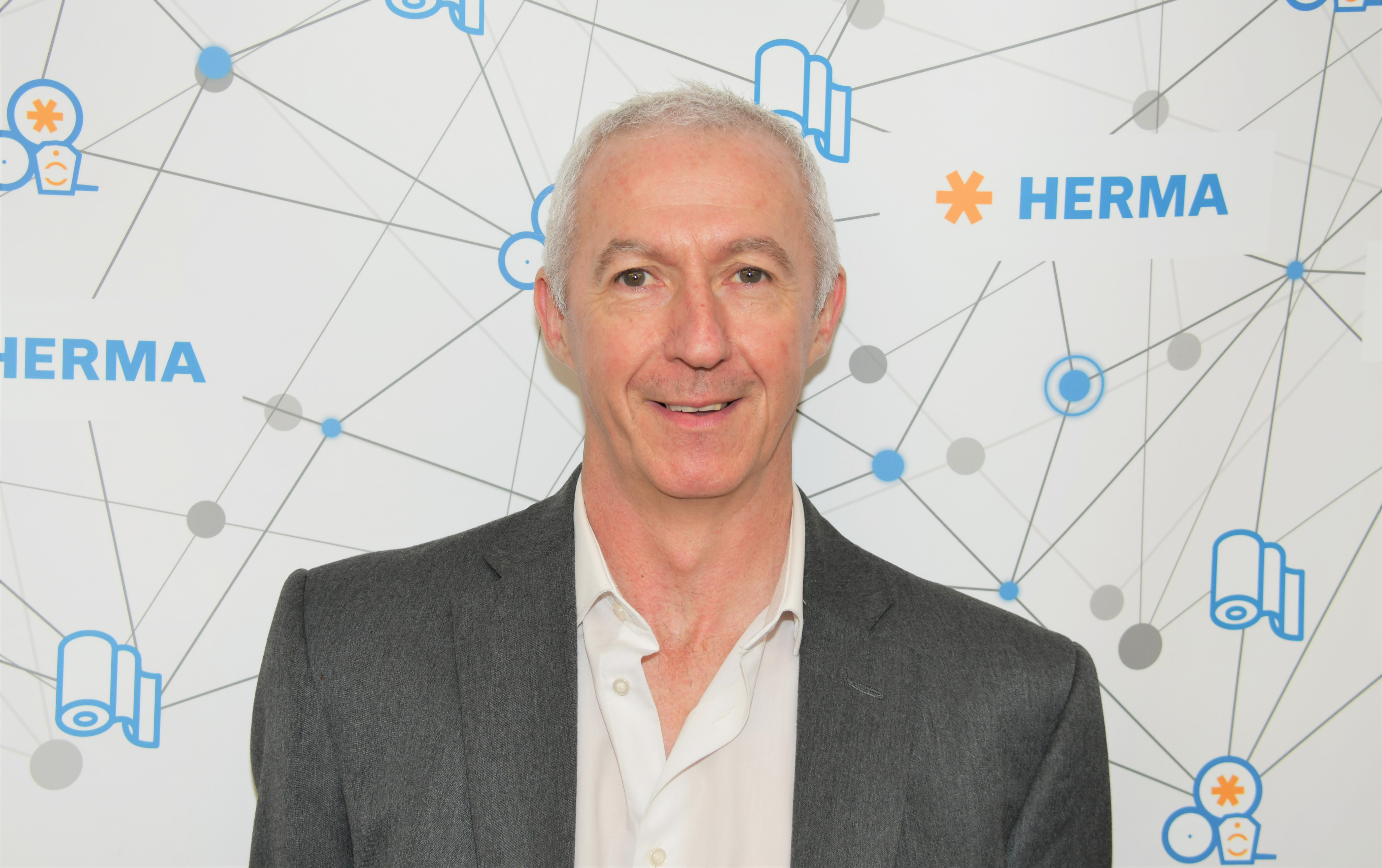 Man in a gray suit and white shirt against a background featuring the HERMA logo and graphic elements.