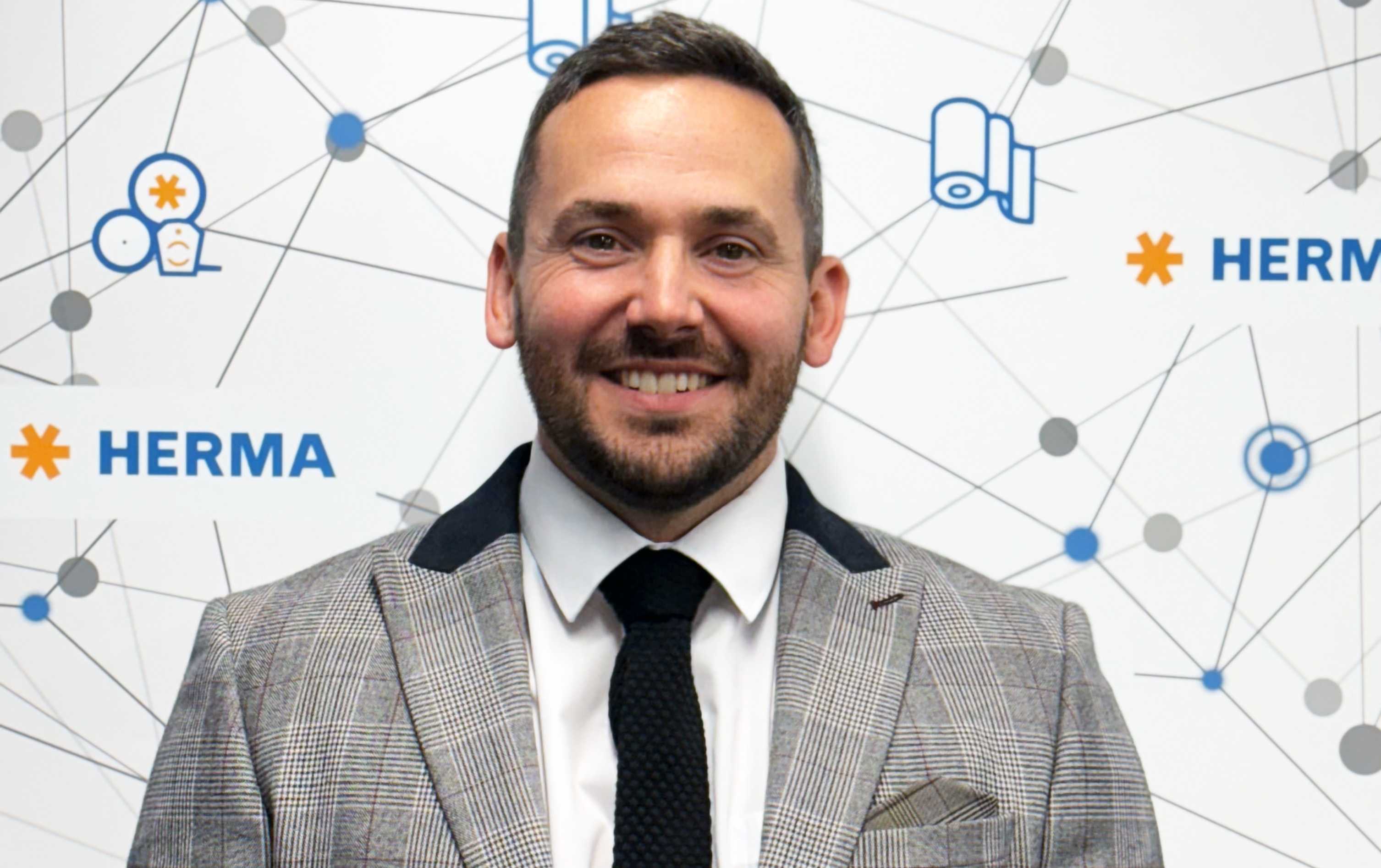 A man in a checked suit and tie smiles against a background featuring the HERMA logo and graphic elements.