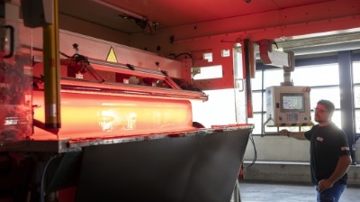 A worker operates a machine that processes a glowing material. Red lighting and technical equipment are visible.